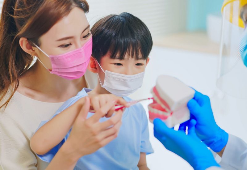 asian dentist with face shield and mask teach boy how use toothbrush brushing teeth use denture model when mother sit by in dental clinic - kid practice it