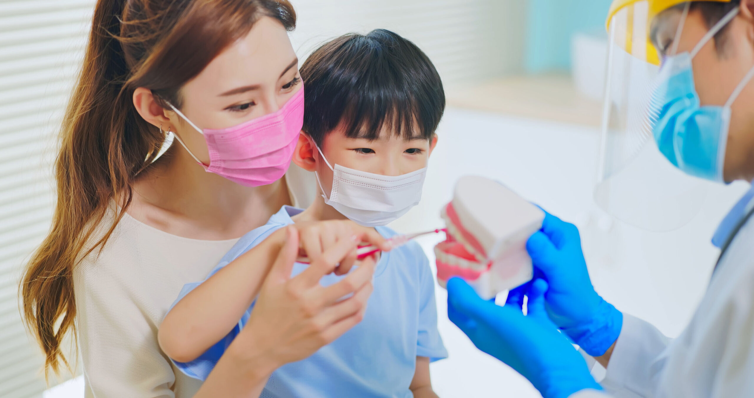 asian dentist with face shield and mask teach boy how use toothbrush brushing teeth use denture model when mother sit by in dental clinic - kid practice it
